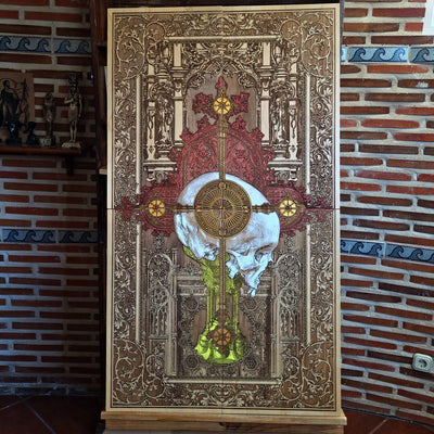 A detailed wooden artwork featuring a skull at the center, overlaid with a cross and intricate gothic architectural elements. The artwork is primarily brown with accents of gold, red, and yellow. 