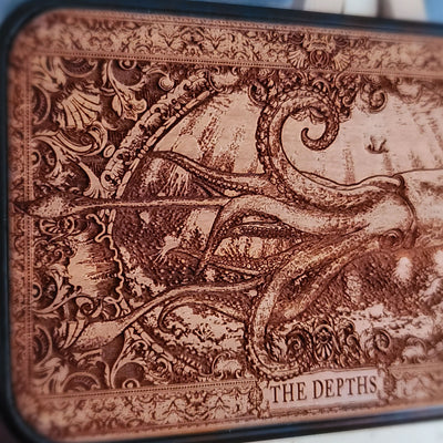 A wood phone case featuring a detailed engraved octopus scene with the text 'THE DEPTHS' at the bottom,