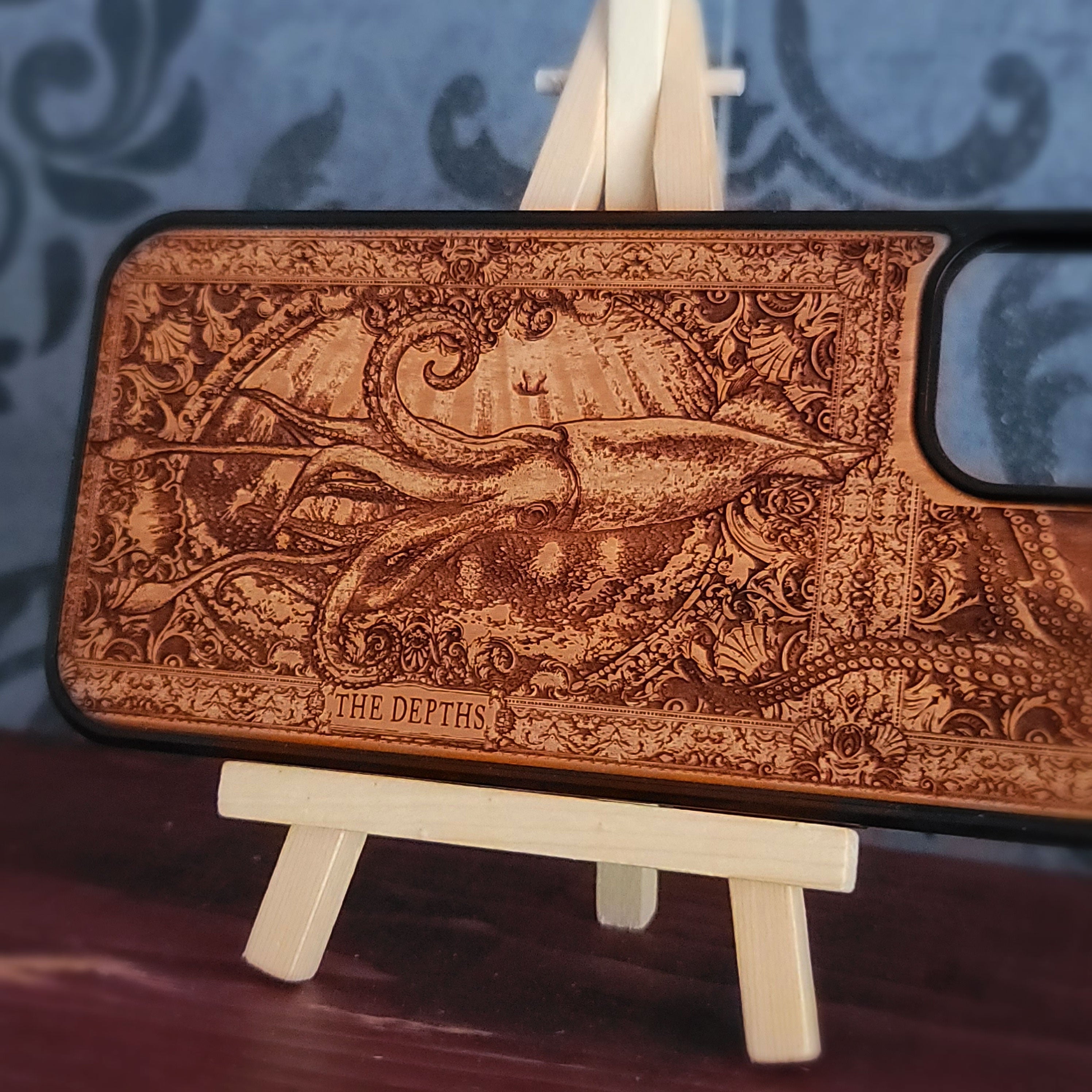 A wooden phone case with an intricate laser-engraved octopus design and the text 'THE DEPTHS' at the bottom, 