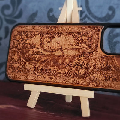 A wooden phone case with an intricate laser-engraved octopus design and the text 'THE DEPTHS' at the bottom, 
