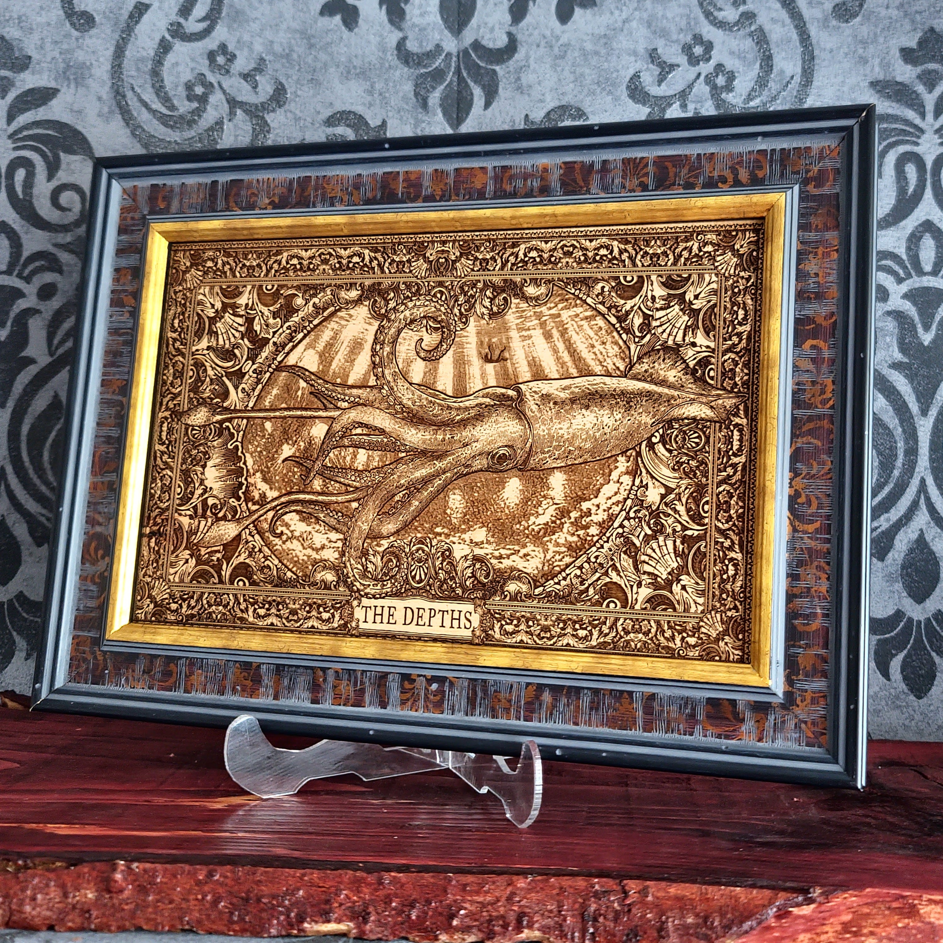 A framed decorative artwork featuring a raised octopus or squid-like marine creature with the words 'THE DEPTHS' at the bottom, displayed on a stand against a patterned wallpaper background.