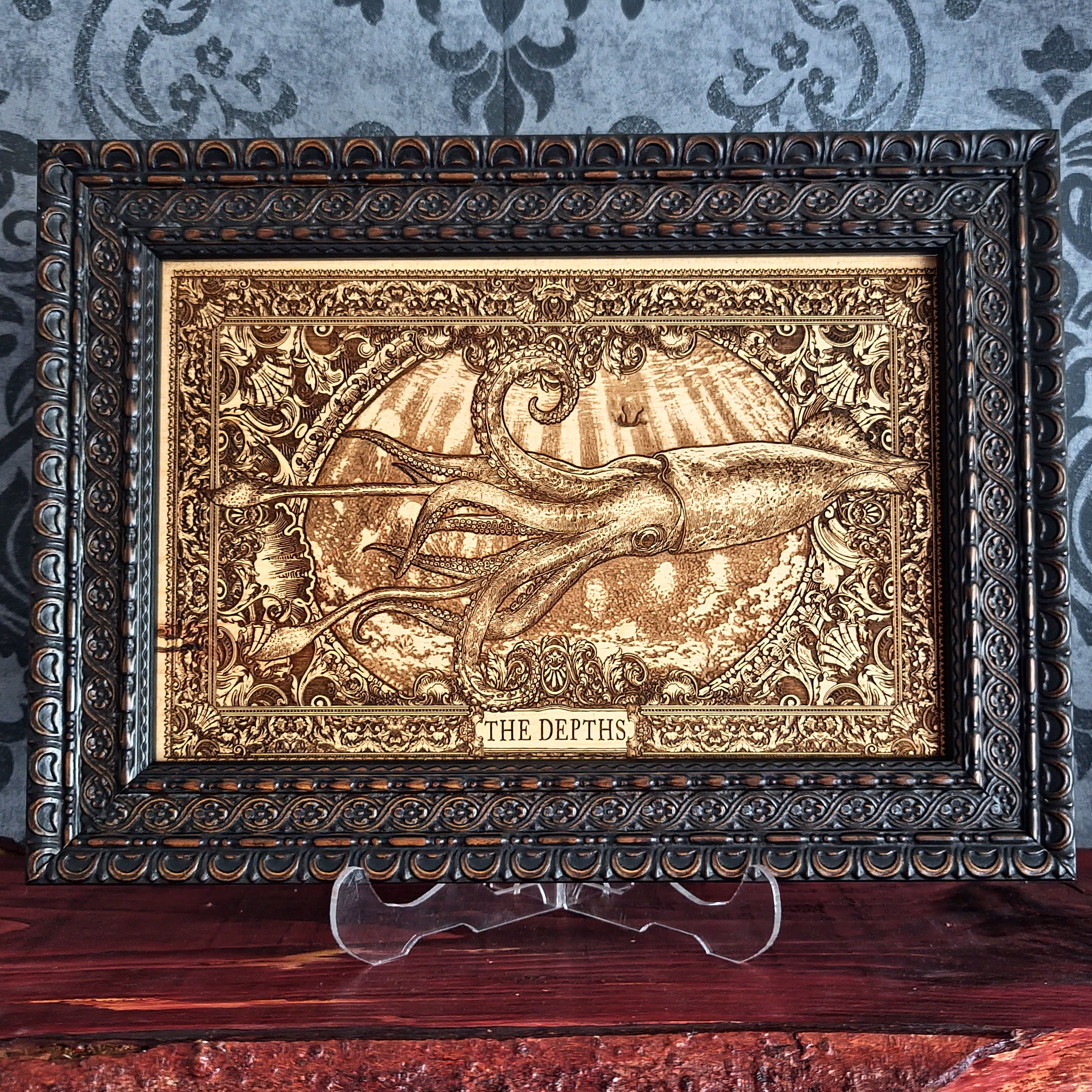 A framed decorative artwork featuring an embossed octopus underwater scene with rays of light and the words 'THE DEPTHS' at the bottom, displayed on a clear stand over a wooden surface against a patterned wallpaper background.