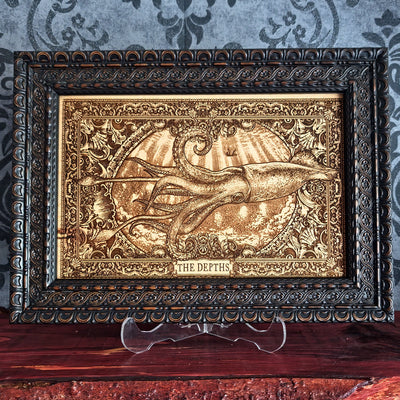 A framed decorative artwork featuring an embossed octopus underwater scene with rays of light and the words 'THE DEPTHS' at the bottom, displayed on a clear stand over a wooden surface against a patterned wallpaper background.