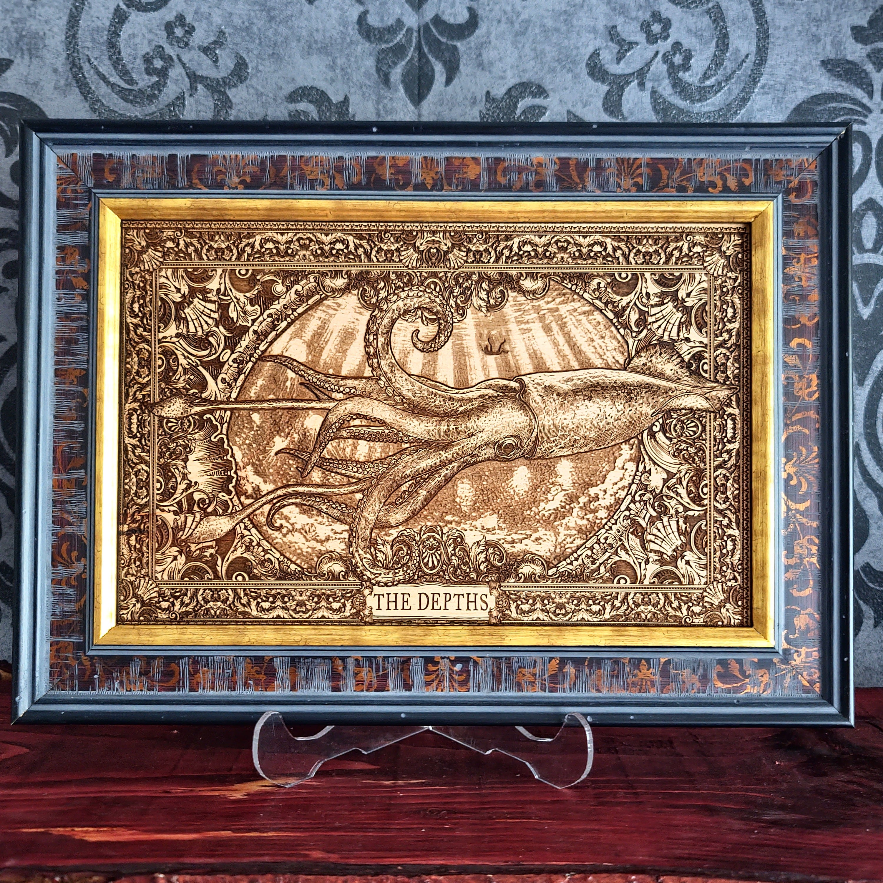 A framed decorative art piece featuring a raised, metallic-looking illustration of a squid or octopus in an ocean scene with the text 'THE DEPTHS' at the bottom. The artwork is set within an ornate gold-toned inner frame and a dark outer frame, displayed on a clear stand against a patterned gray wall and wooden surface.