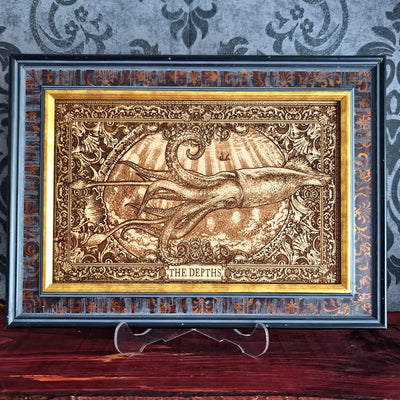 A framed decorative art piece featuring a raised, metallic-looking illustration of a squid or octopus in an ocean scene with the text 'THE DEPTHS' at the bottom. The artwork is set within an ornate gold-toned inner frame and a dark outer frame, displayed on a clear stand against a patterned gray wall and wooden surface.
