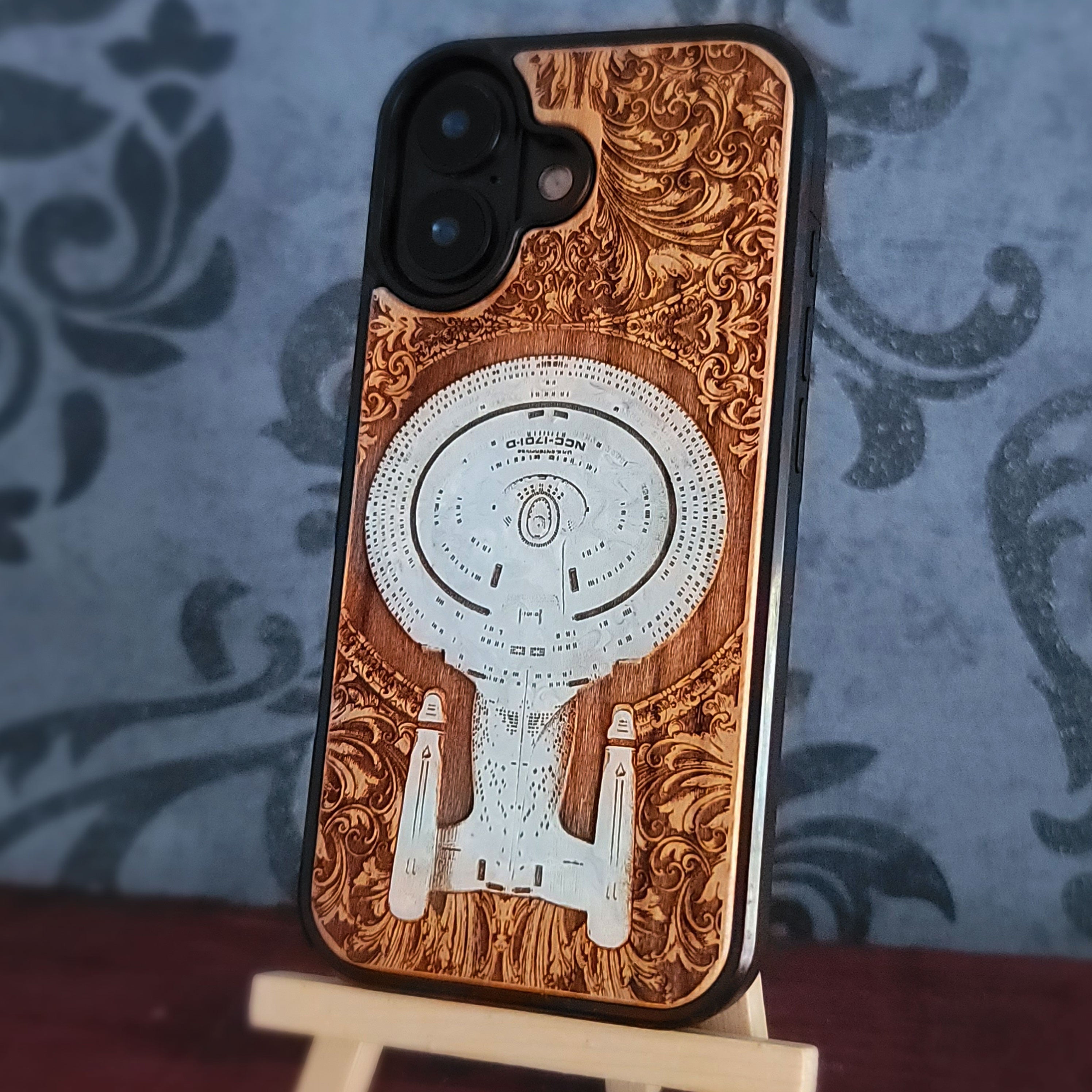 Enterprise NCC-1701-D hand-painted cherrywood phone case, three-quarter view displayed on a tabletop easel