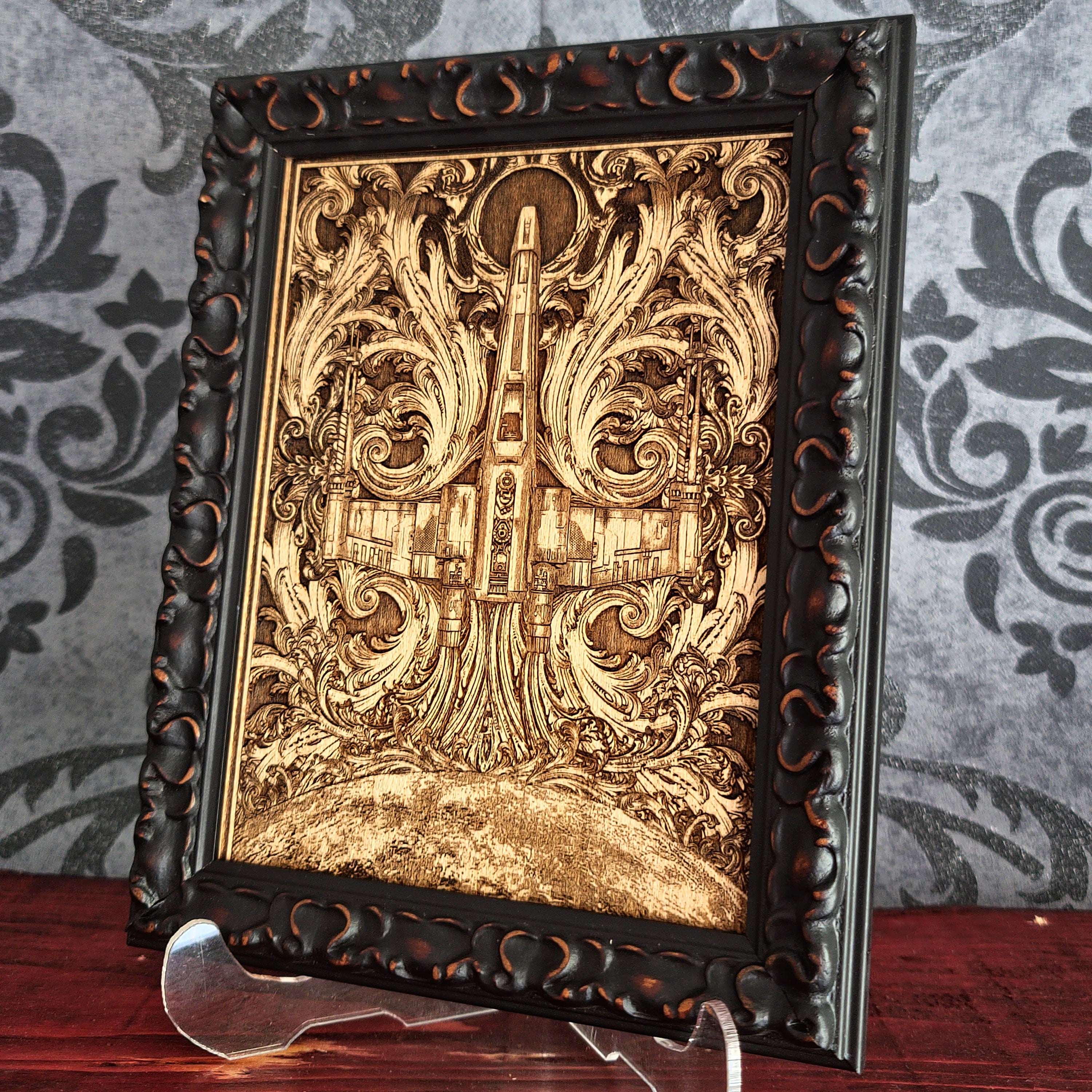 Wooden laser-cut artwork featuring a detailed depiction of a spaceship, surrounded by ornate, swirling floral patterns. The artwork is framed and has a natural wood color, with intricate details and a vintage aesthetic.