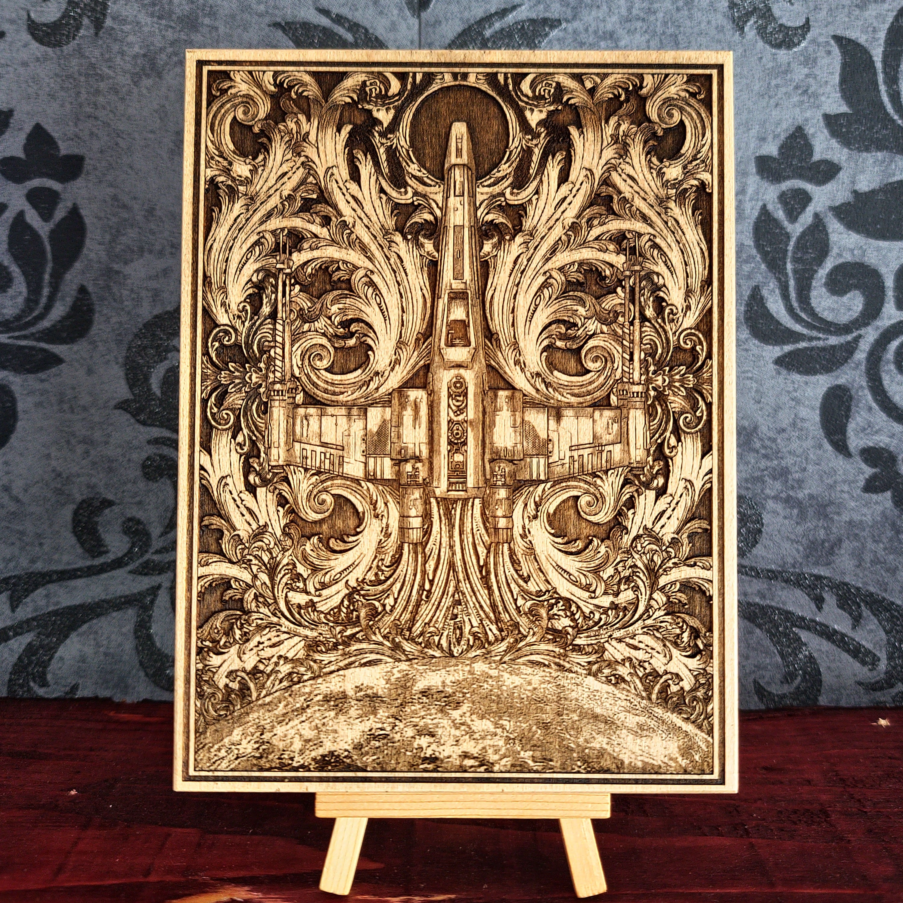 Wooden laser-cut artwork featuring a detailed depiction of a spaceship, surrounded by ornate, swirling floral patterns. The artwork is framed and has a natural wood color, with intricate details and a vintage aesthetic.
