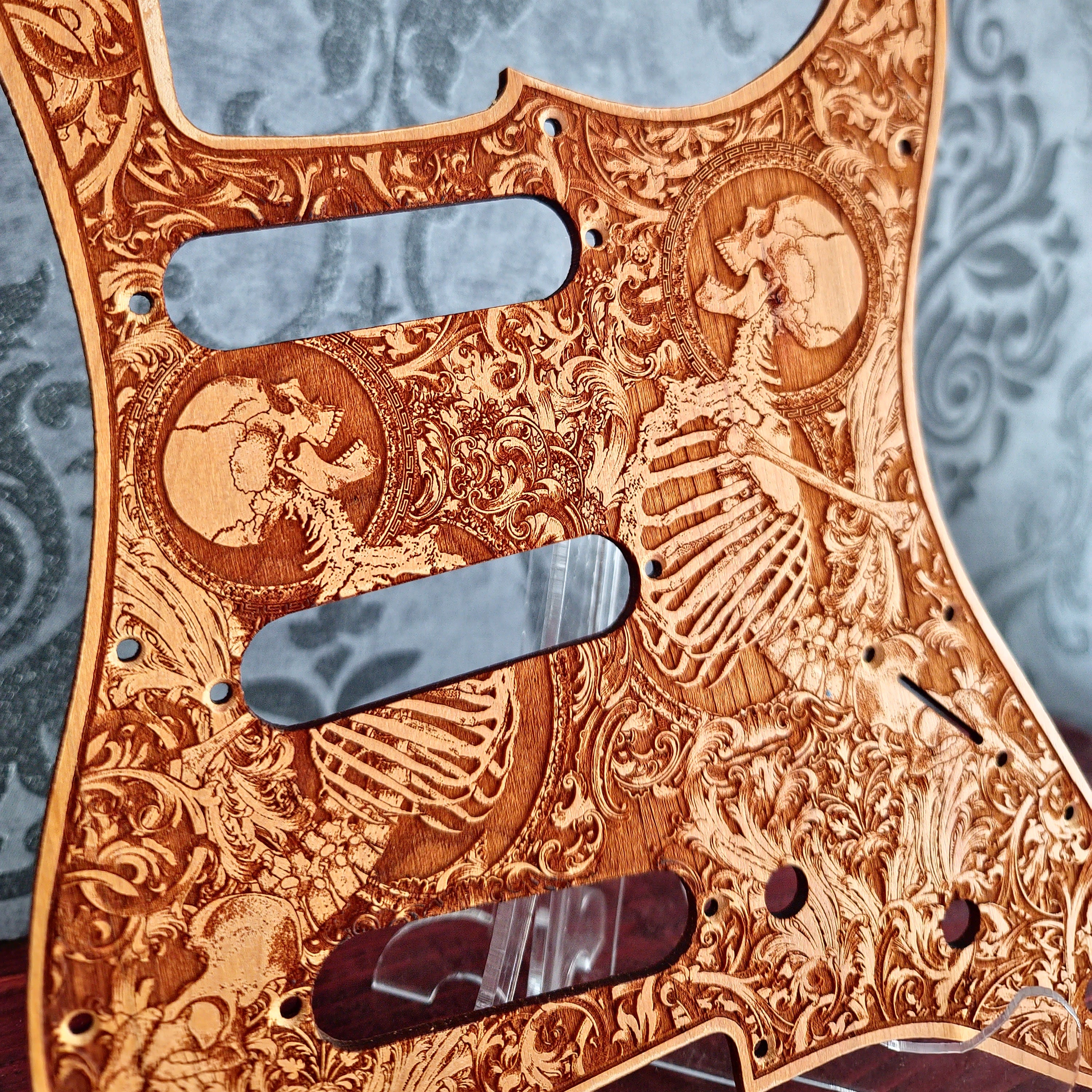 A close-up of a guitar pickguard featuring an intricate laser-engraved skeleton and scrollwork design on a natural wood background, with three dark metallic cutouts.