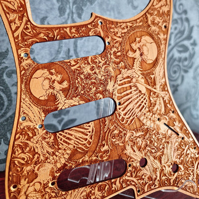 A close-up of a guitar pickguard featuring an intricate laser-engraved skeleton and scrollwork design on a natural wood background, with three dark metallic cutouts.