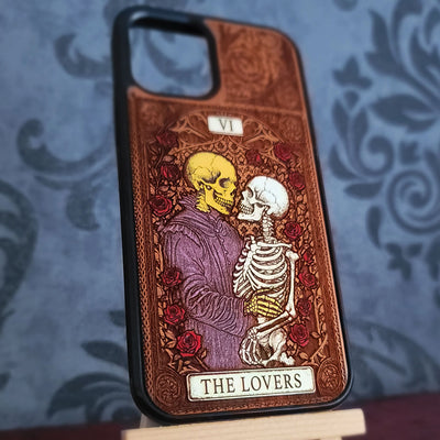 A phone case featuring a tarot card design titled 'THE LOVERS' (VI). The design depicts a yellow-skinned lover kissing a white-skinned lover, surrounded by red roses and a purple robe, set against a brown ornate background.