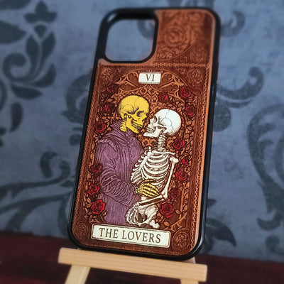 A phone case featuring the 'The Lovers' tarot card design, displayed on a small wooden stand against a blue damask patterned background. The design depicts a kissing yellow and white skeleton surrounded by red roses on a brown textured background.