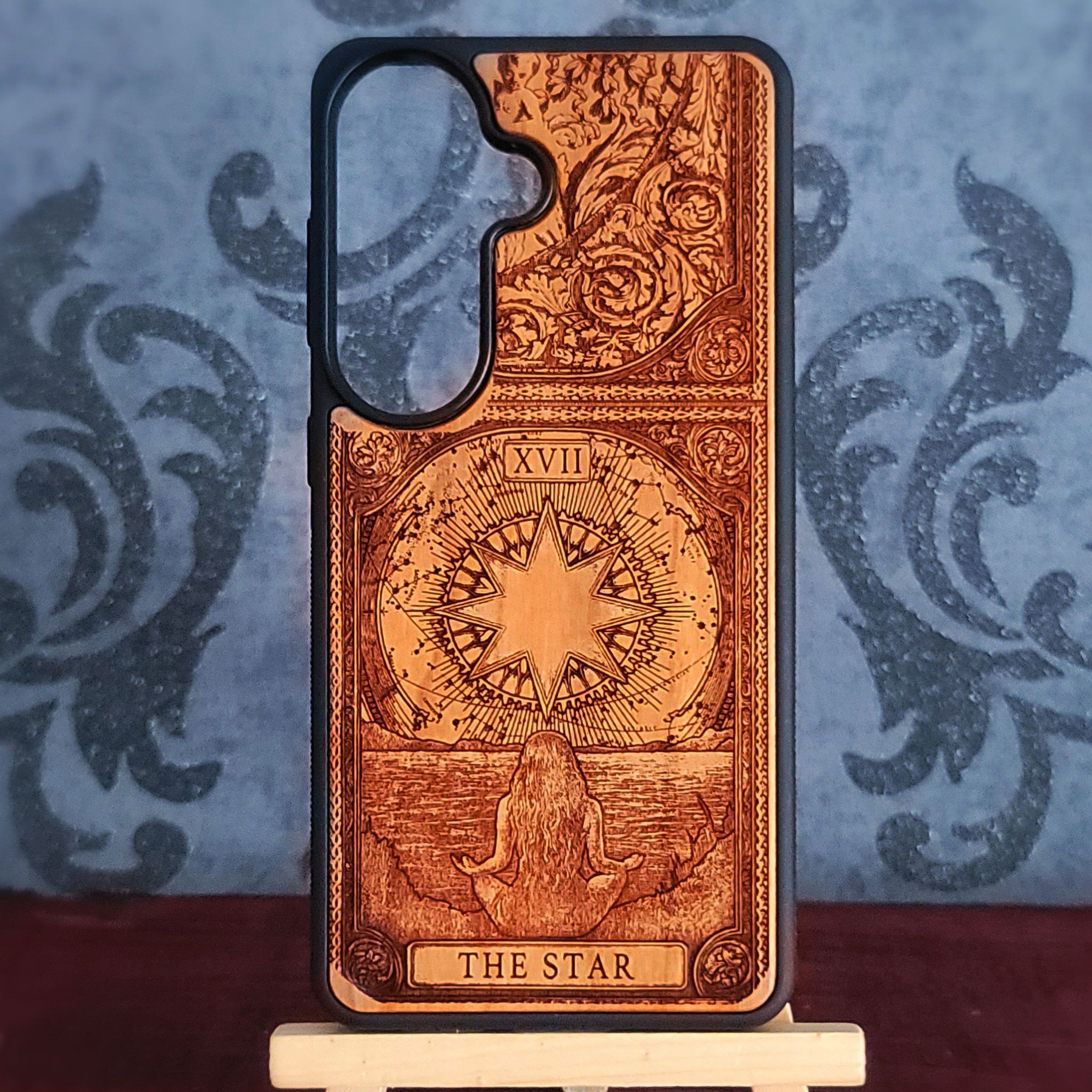 A phone case featuring a detailed, laser-engraved Tarot card design of 'The Star' on a warm brown wood-grain finish, displayed on a small white stand against a blue patterned backdrop.