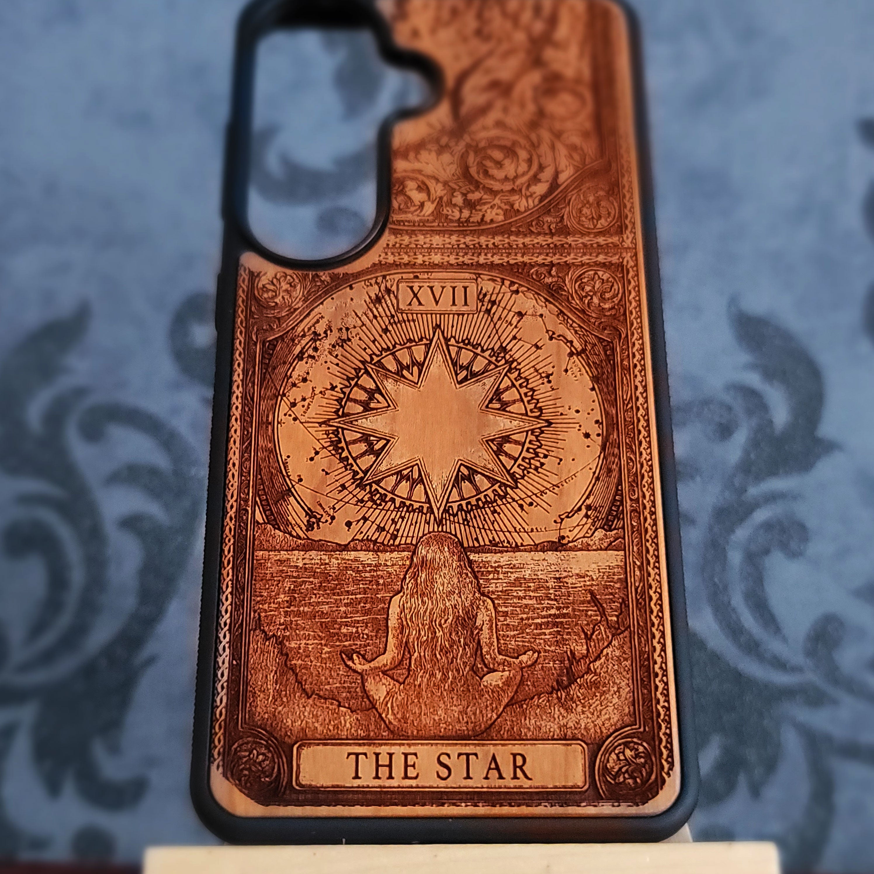 A wooden tarot card phone case featuring 'The Star' card design, displayed on a blue damask-patterned surface.