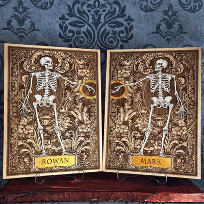 Two personalized gothic-style skeleton cards displayed side-by-side on small clear stands. The cards feature detailed ornate brown and gold backgrounds with white skeleton illustrations, one labeled 'ROWAN' and the other 'MARK'.
