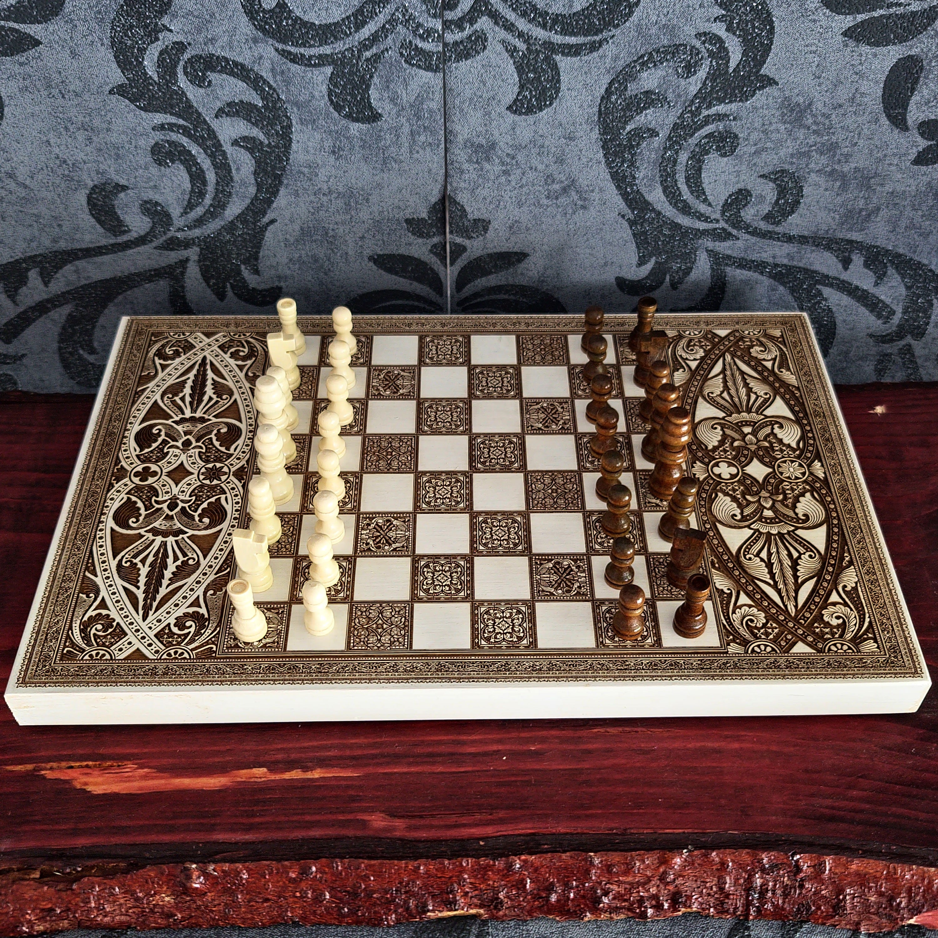 An ornate decorative chess set displayed on a dark wooden table, featuring carved ivory and dark brown pieces with intricate geometric and floral patterns on the board and pieces, set against a gray damask wallpaper background.