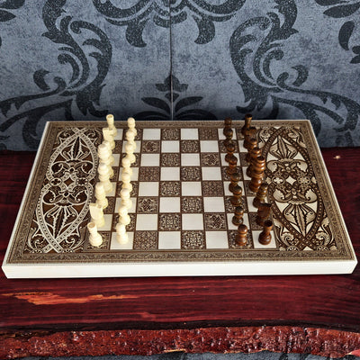 An ornate decorative chess set displayed on a dark wooden table, featuring carved ivory and dark brown pieces with intricate geometric and floral patterns on the board and pieces, set against a gray damask wallpaper background.