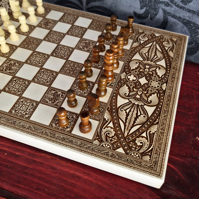 An ornate chessboard with carved ivory and dark brown squares, decorated with intricate floral engravings along the right edge. Wooden chess pieces are arranged on the board, with the white pieces partially visible in the upper left and the dark brown pieces in the foreground, set on a dark wooden table.