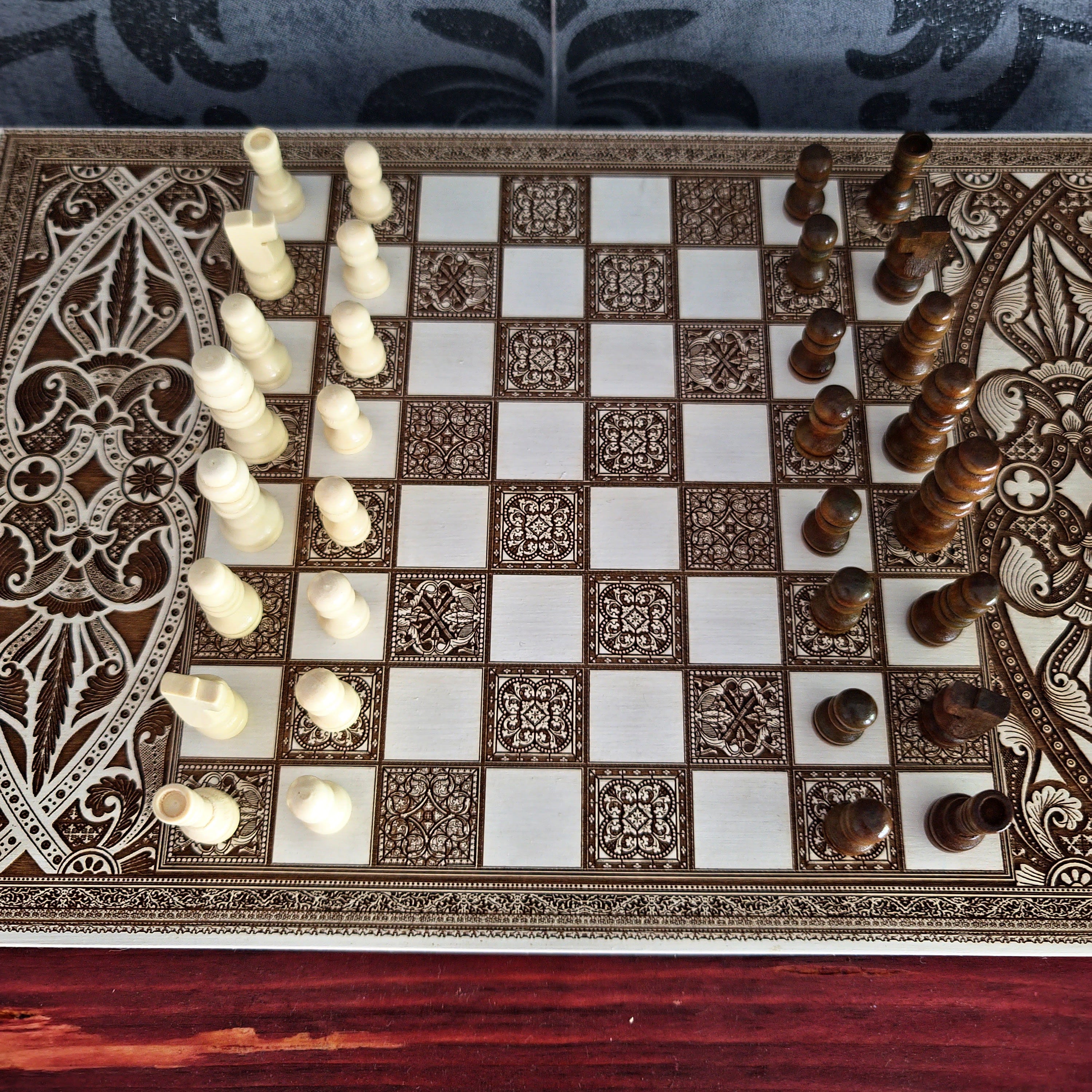 An ornate rectangular chessboard with intricate carved or inlaid patterns around the border and in the center of each square. The board has alternating cream and dark brown checkers, with white and dark brown chess pieces arranged on it.