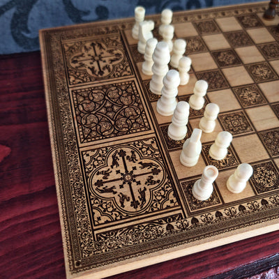 A decorative wooden chessboard with ornate dark engraved patterns and white chess pieces arranged on one diagonal, photographed on a dark wooden surface.
