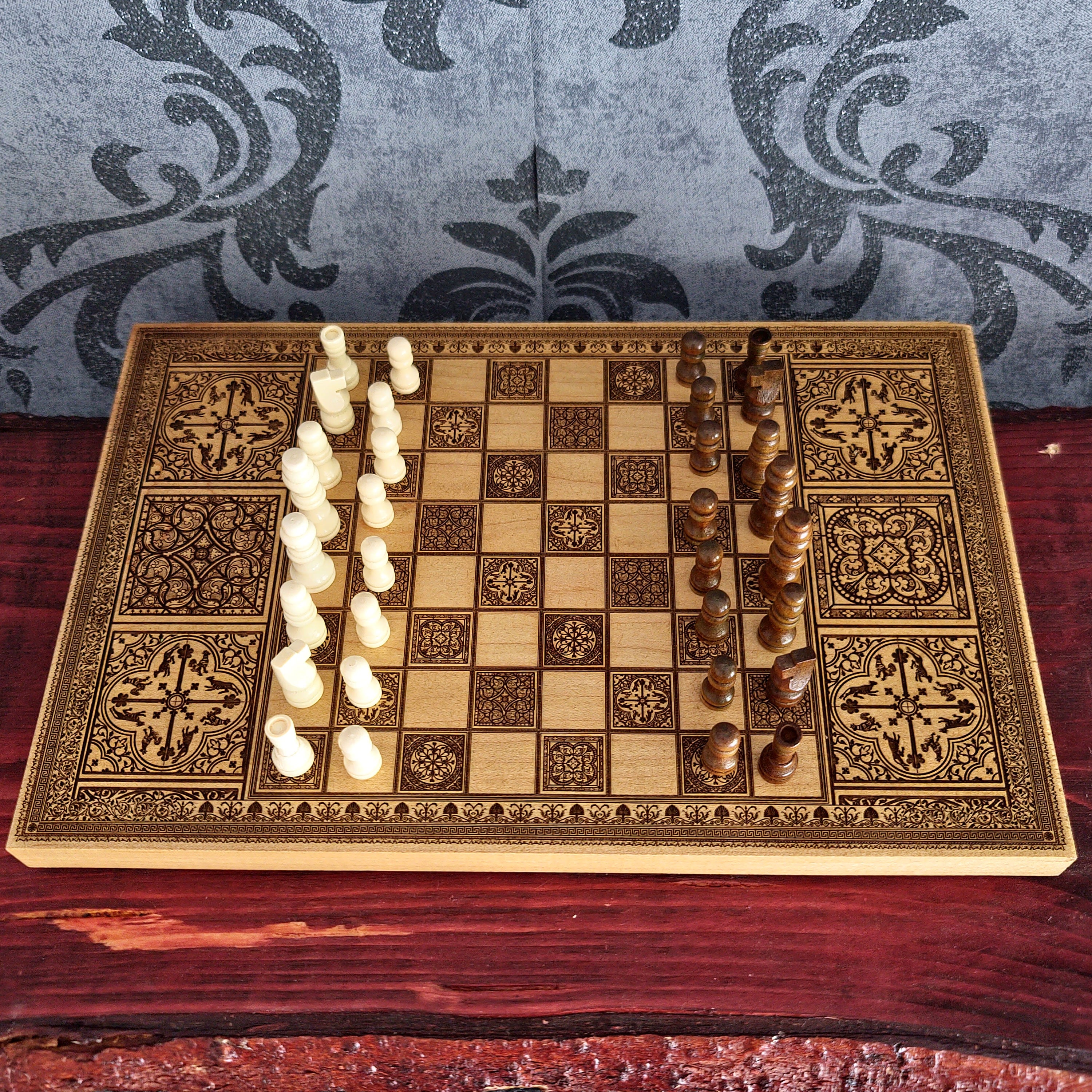 An ornate wooden chessboard with intricate geometric and floral engravings, set on a dark red wooden surface. The board is divided down the middle, with one side featuring off-white/cream chess pieces and the other side featuring dark brown wooden chess pieces.