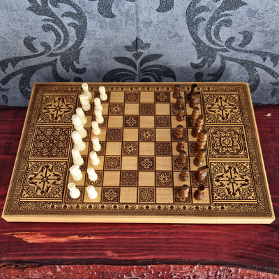 An ornate wooden chessboard with intricate geometric and floral engravings, set on a dark red wooden surface. The board is divided down the middle, with one side featuring off-white/cream chess pieces and the other side featuring dark brown wooden chess pieces.