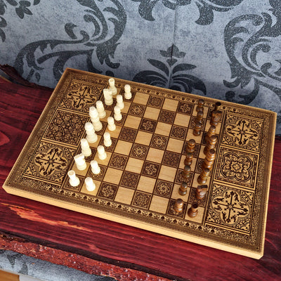 An ornate rectangular wooden chessboard with engraved decorative patterns, set on a dark red wooden surface. The board has cream-colored chess pieces on one side and dark brown chess pieces on the other, arranged for play.