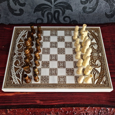An ornate rectangular chessboard with carved decorative borders, shown with wooden and ivory chess pieces arranged 