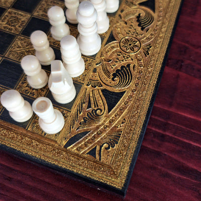 An ornate chess set with carved ivory chess pieces and a black board featuring intricate gold filigree borders, set 