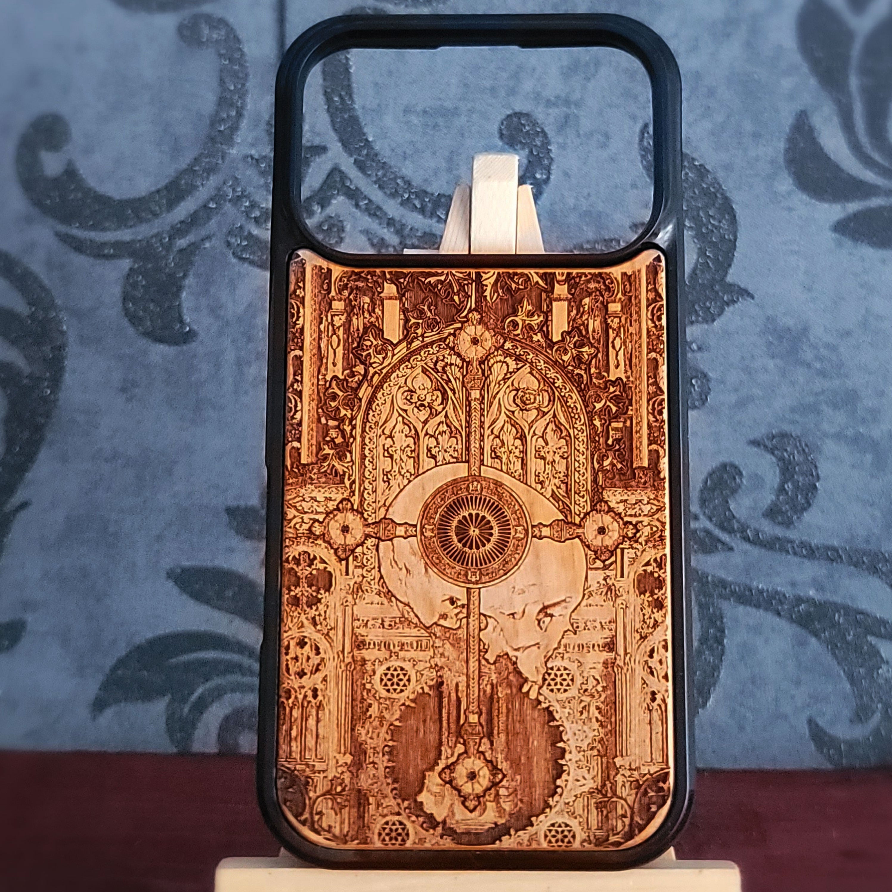 A wooden phone case with a black bumper frame, featuring an intricate laser-burned gothic design of a harp and ornate arches. The case is displayed on a small stand against a blue damask-patterned background.