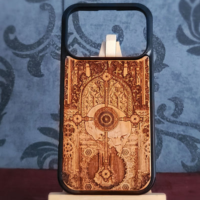 A wooden phone case with a black bumper frame, featuring an intricate laser-burned gothic design of a harp and ornate arches. The case is displayed on a small stand against a blue damask-patterned background.