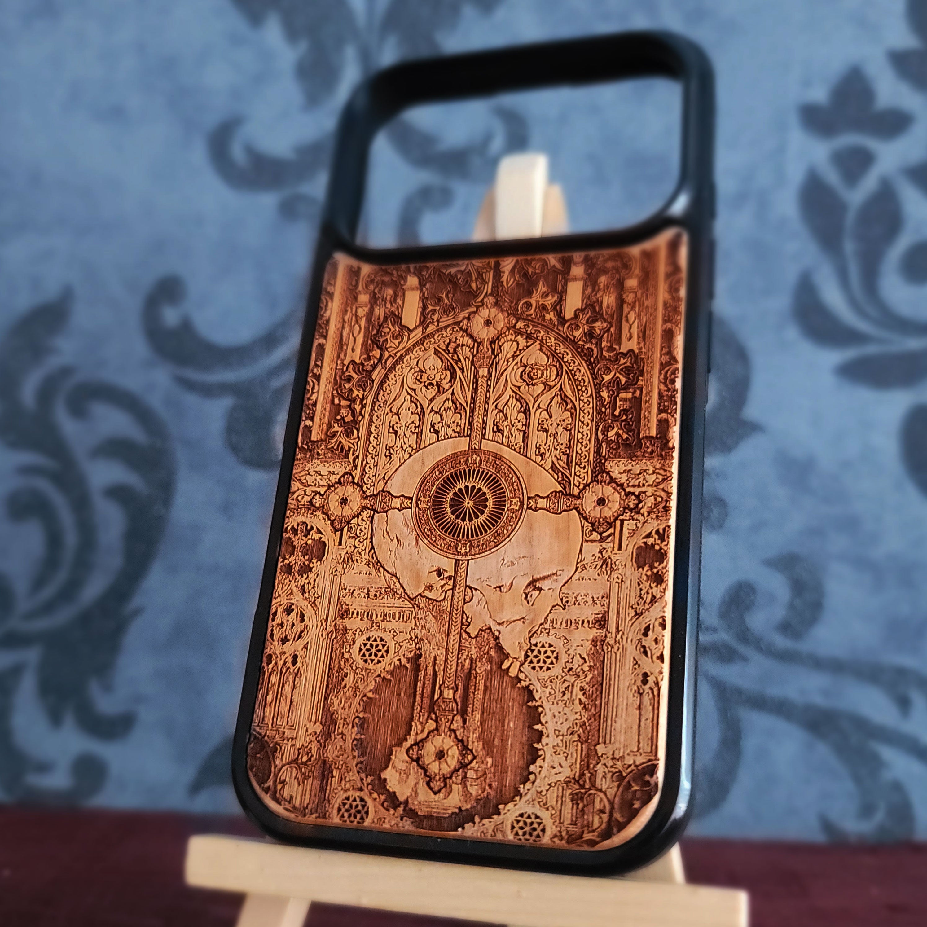 A wooden phone case with a black bumper frame, featuring an intricate laser-burned gothic mandolin design, displayed on a small wooden stand against a blue damask patterned background.