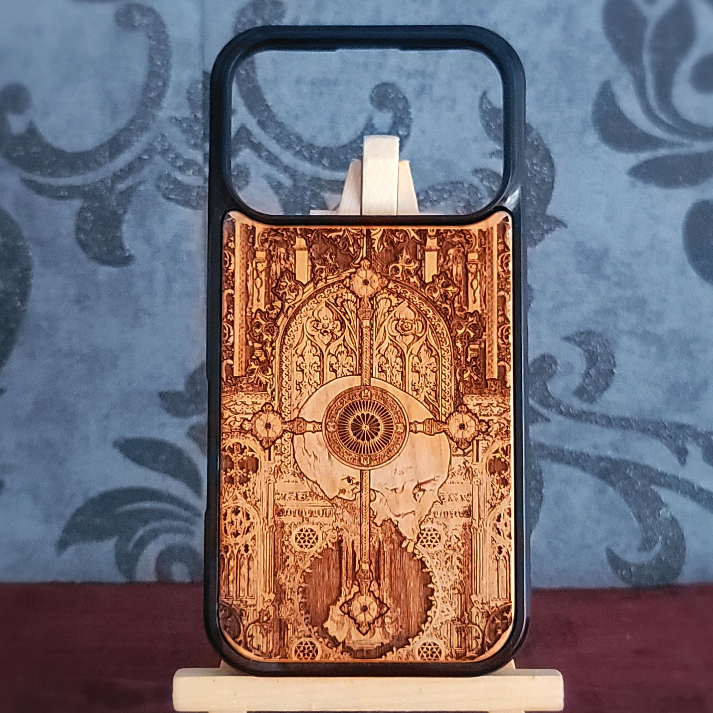 A wooden phone case with an intricate, hand-burned gothic arch and mandala design, displayed on a small stand against a blue patterned wallpaper background.