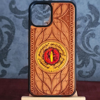A wooden phone case with an embossed Elvish pattern and a central red and yellow Eye of Sauron design, displayed on a small stand against a patterned blue-gray background.