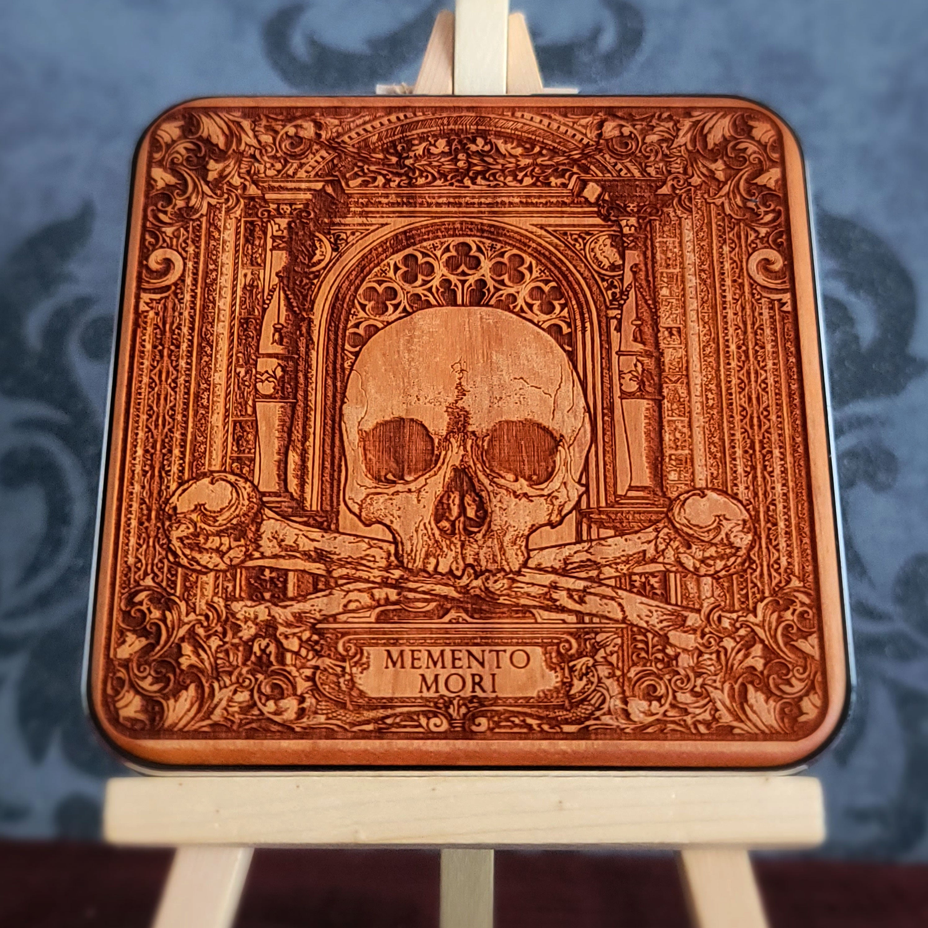 A wooden WIRELESS charger pad with a laser-etched design of a skull, crossed bones, and an ornate archway. The text "Memento Mori" is inscribed below the skull.