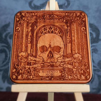 A wooden WIRELESS charger pad with a laser-etched design of a skull, crossed bones, and an ornate archway. The text "Memento Mori" is inscribed below the skull.