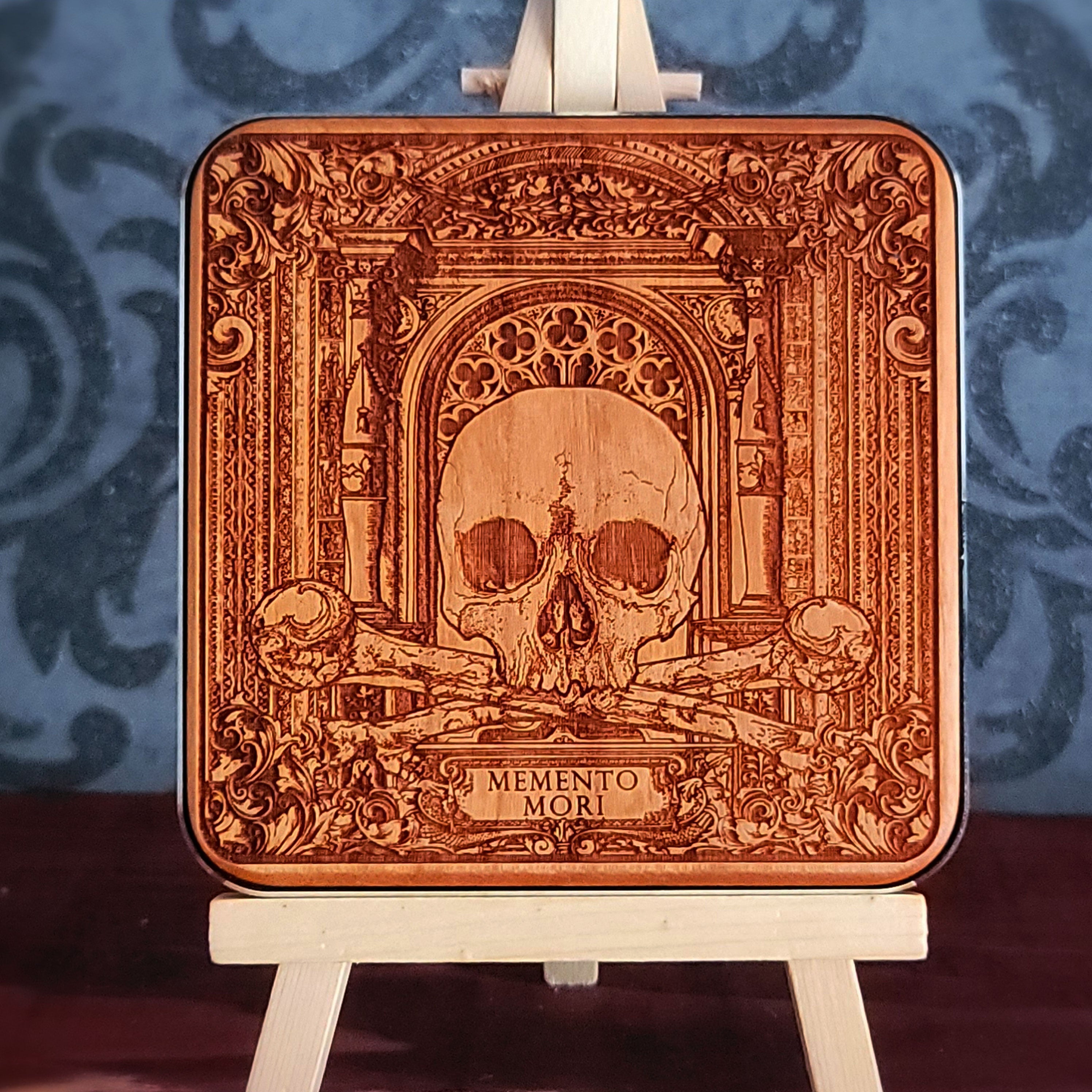 A wooden WIRELESS charger pad with a laser-etched design of a skull, crossed bones, and an ornate archway. The text "Memento Mori" is inscribed below the skull.