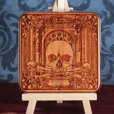 A wooden WIRELESS charger pad with a laser-etched design of a skull, crossed bones, and an ornate archway. The text "Memento Mori" is inscribed below the skull.