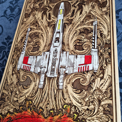 A close-up of a decorative wooden panel or artwork featuring a white X-wing style spacecraft over ornate swirling wood-carved patterns, with red accents and a blue patterned fabric visible at the sides.